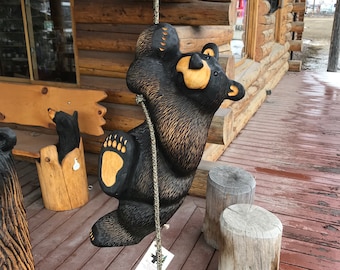 May include: A black wooden bear sculpture climbing a rope. The bear has a light brown belly and paws. The sculpture is hanging on a wooden porch. Other wooden bear carvings and tree stumps are also visible.