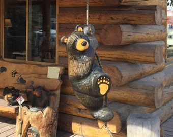 May include: A carved wooden bear sculpture hanging from a rope, with a log cabin in the background. The bear is brown and black with yellow accents and is climbing up the rope.