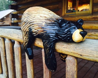 May include: A carved wooden bear sculpture with black and brown paint, resting on a wooden railing. The bear has a yellow nose and is positioned as if it is hanging off the railing.