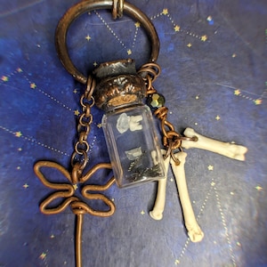 May include: A copper necklace with a small glass vial filled with white and black objects. The vial is attached to a copper chain with a copper dragonfly charm and two bone charms.