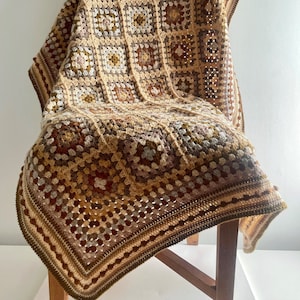 Puede incluir: Una manta de ganchillo en tonos marrones, beige y crema con un patrón de cuadrados de la abuela. La manta está drapeada sobre una silla de madera.