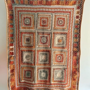 Può includere: Una coperta quadrata all'uncinetto fatta a mano nei toni caldi dell'arancione, del rosso, del grigio e del crema. La coperta presenta un motivo ripetuto di quadrati dentro quadrati, con un bordo dettagliato. È appesa a un filo con mollette bianche.