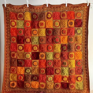 May include: A handmade crochet blanket in a patchwork design, featuring squares in shades of orange, red, and yellow. The blanket has a detailed border and is hung with white clips. Autumn leaves are arranged at the bottom.