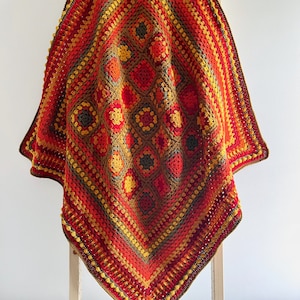 May include: A handmade crochet blanket in red, orange, yellow, and brown. The blanket has a geometric pattern with a central diamond design and a textured border. It is draped over a wooden ladder, showcasing the vibrant colours.