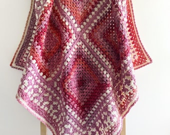 May include: A crocheted blanket with a diamond pattern in shades of pink, red, and white. The blanket is draped over a wooden stand.