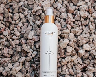 May include: A white bottle of "Cheeky Curls" curl volumizer with a gold cap and spray nozzle. The bottle has black text that reads "Curl Volumizer" and "Rose Water Hair Elixir". The bottle is 8 oz / 236 ml.