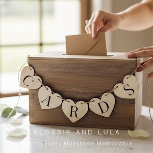 May include: A brown wooden card box with a slot, decorated with a heart-shaped garland spelling "CARDS." A hand is posting a card. The text "FLOSSIE AND LULA" is visible at the bottom, with the tagline.