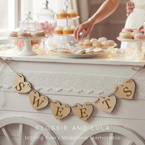 May include: A dessert table with cupcakes, cookies, and candies. A wooden heart banner spells "SWEETS". The table is white with lace trim. The text "FLOSSIE AND LULA Making Every Milestone Memorable" is at the bottom.