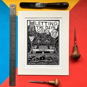 Op de afbeelding: Zwart-witte linosnede van een auto die over een weg rijdt met een huis op de achtergrond. De auto heeft het woord "TLKNGHDS" op het kenteken. De tekst op de print luidt "Letting the days go by, Same as it ever was, Once in a life time, water flowing underground".