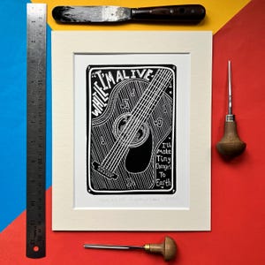 Könnte beinhalten: Schwarz-Weiß-Linolschnitt mit einer Akustikgitarre und Noten. Der Druck enthält den Text "While I'm Alive" und "I'll Make Tiny Changes To Earth". Das Kunstwerk ist in einem weißen Passepartout gerahmt.