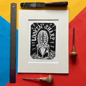 Peut inclure: Gravure sur linoléum en noir et blanc d'un cactus portant un smoking avec le texte "Lookin' Sharp" dans une police décorative.