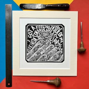 May include: Black and white linocut print in a cream-coloured mount. The print features a rose above stylised waves, with the text "I got a kinda strange feeling inside of me like a storm sea." The print is framed.