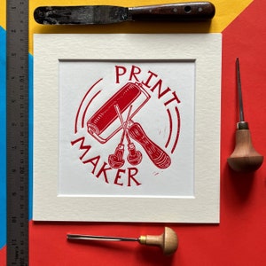 Könnte beinhalten: Ein roter Druck eines Gravurwerkzeug-Sets mit einer Rolle, zwei Werkzeugen und dem Text "PRINT MAKER" in einem Kreis.