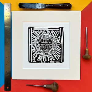 May include: Black and white art print with the text "Hey, Have You Been Trying to Meet You?" in a decorative frame. The print is matted and framed in a cream-coloured mat. A ruler, a palette knife, and two carving tools are also visible.
