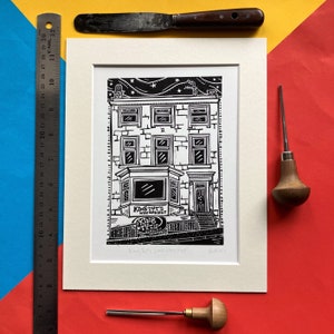 May include: A black and white linocut print of a building with a sign that reads "King Tut's Wah Wah Hut". The print is framed in a white mount and has a white background. The print is titled "King Tut's Wah Wah Hut" and is dated "Feb 2014".