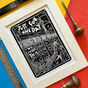 May include: Black and white linocut print of a cityscape with a tall tower and the text "Just for one day" and "We can beat them". The print is framed in a white mat.