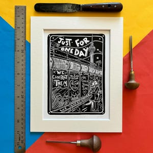 May include: Black and white linocut print of a couple walking down a street in Berlin, Germany. The print features the text "Just for one day" and "We can beat them".