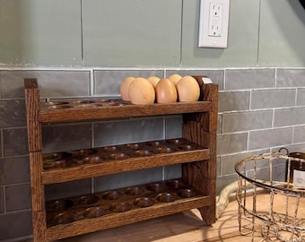 May include: A three-tiered wooden egg rack, stained a warm brown, holds several brown eggs. The rack has carved details and is designed to store eggs. The rack is on a wooden surface.