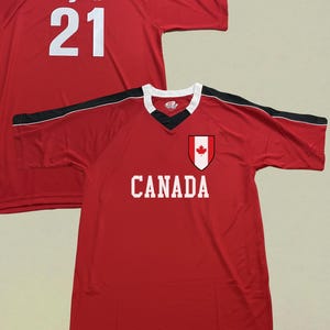 May include: Red jersey with black and white detailing. The jersey has the name "Ryba" and the number "21" in white on the back. The front of the jersey shows the word "CANADA" and a Canadian flag emblem.