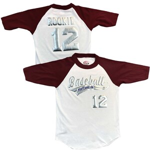 May include: White baseball jersey with maroon sleeves. The jersey features the word "Baseball" above "Portland, OR" and the number "12" in silver. The back of the jersey has the word "ROOKIE" and the number "12" in silver.