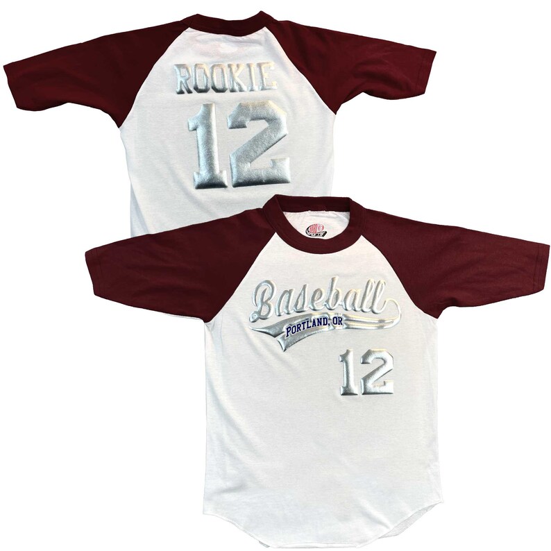 May include: White baseball jersey with maroon sleeves. The jersey features the word "Baseball" above "Portland, OR" and the number "12" in silver. The back of the jersey has the word "ROOKIE" and the number "12" in silver.