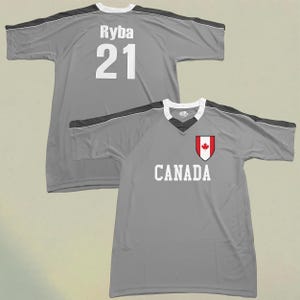 May include: Two grey athletic jerseys. One jersey has the name "Ryba" and the number "21" in white. The other jersey has the word "CANADA" in white, and a red and white Canadian flag emblem.