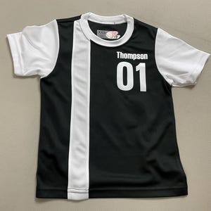 May include: Black and white athletic jersey with the name "Thompson" and the number "01" in white. The jersey has white sleeves and a white vertical stripe down the front. The jersey is a size XXS.