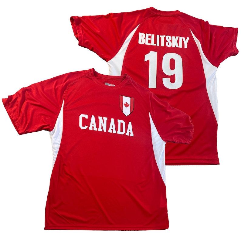 May include: Red Canada soccer jersey with white accents. The front displays "CANADA" in white block letters and a maple leaf crest. The back features "BELITSKIY" and the number "19" in white. The jersey has short sleeves.