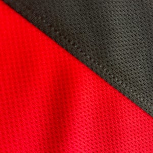May include: Close-up of a red and black athletic fabric. The red fabric has a textured, perforated pattern. The black fabric is a darker shade and is also textured. The two fabrics are sewn together diagonally.