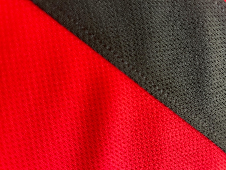 May include: Close-up of a red and black athletic fabric. The red fabric has a textured, perforated pattern. The black fabric is a darker shade and is also textured. The two fabrics are sewn together diagonally.