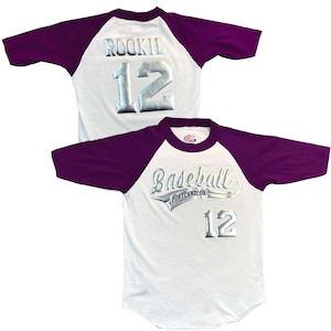 May include: White baseball jersey with purple sleeves. The jersey features the word "ROOKIE" and the number "12" in silver lettering on the back. The front of the jersey says "Baseball Portland, OR" and the number "12" in silver lettering.