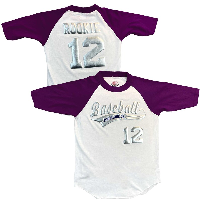 May include: White baseball jersey with purple sleeves. The jersey features the word "ROOKIE" and the number "12" in silver lettering on the back. The front of the jersey says "Baseball Portland, OR" and the number "12" in silver lettering.