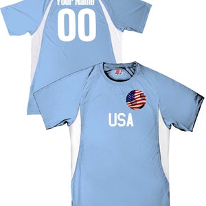May include: Light blue soccer jersey with white accents. The back of the jersey has the text "Your Name" and the number "00". The front of the jersey has the text "USA" and a soccer ball with an American flag.