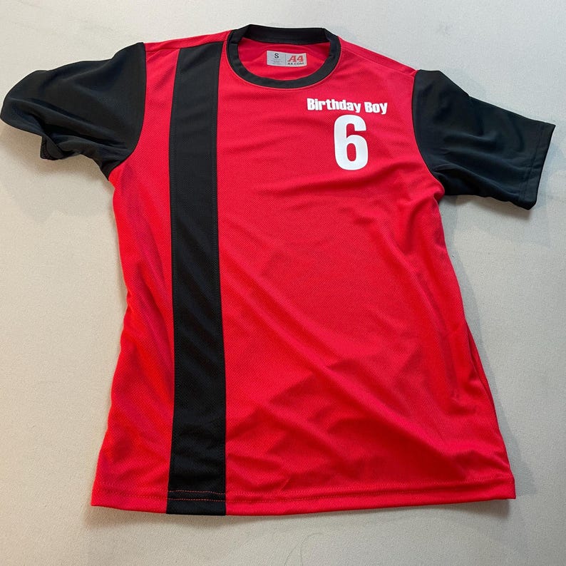 May include: Red and black short-sleeved jersey with the text "Birthday Boy 6". The jersey has a black vertical stripe on the left side and black sleeves. The shirt is made of a breathable, athletic material.