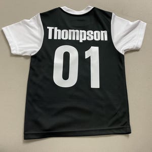 May include: Black and white athletic jersey with the name "Thompson" and the number "01" printed on the back. The jersey has white sleeves and a white collar. The jersey is made of a breathable, mesh-like material.