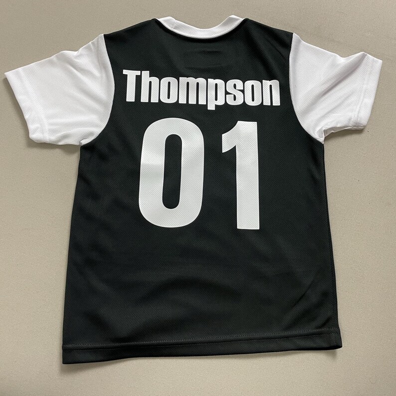May include: Black and white athletic jersey with the name "Thompson" and the number "01" printed on the back. The jersey has white sleeves and a white collar. The jersey is made of a breathable, mesh-like material.