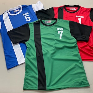 May include: Three athletic jerseys in blue, green, and red. The blue jersey has white accents and the number 5. The green jersey has a black stripe and the number 7. The red jersey has black sleeves and the number 1. Each jersey has the name of a country.