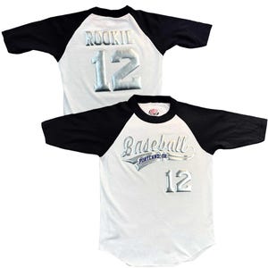 May include: White baseball jersey with black sleeves. The jersey features the word "ROOKIE" and the number "12" in silver lettering on the back. The front of the jersey has the word "Baseball" and the number "12" in silver lettering.