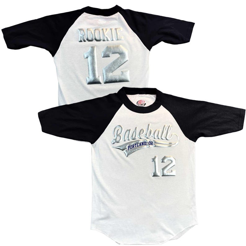 May include: White baseball jersey with black sleeves. The jersey features the word "ROOKIE" and the number "12" in silver lettering on the back. The front of the jersey has the word "Baseball" and the number "12" in silver lettering.