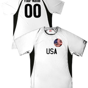 May include: White soccer jersey with black side panels. The back of the jersey has "Your Name" and "00" printed on it. The front of the jersey has "USA" printed on it with a soccer ball with an American flag design.