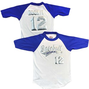 May include: Two baseball jerseys. One white jersey with blue sleeves has "Baseball Portland, OR" and the number "12" in silver. The other jersey has "Rookie 12" in silver on the back.