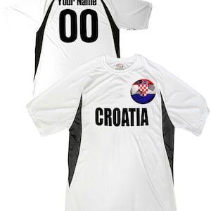 Puede incluir: Camiseta de f&uacute;tbol blanca con detalles en negro y gris. La camiseta tiene un bal&oacute;n de f&uacute;tbol con la bandera croata y la palabra "CROATIA" impresa en la parte delantera. La parte posterior de la camiseta tiene un n&uacute;mero negro "00" y el texto "Your Name" impreso.