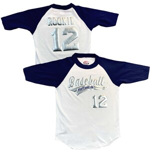 May include: White baseball jersey with navy blue sleeves. The jersey features the word "ROOKIE" and the number "12" in silver lettering on the back. The front of the jersey says "Baseball PORTLAND, OR" and the number "12".