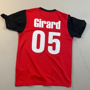 May include: Red jersey with black sleeves and a black collar. The name "Girard" and the number "05" are printed in white on the back of the jersey. The jersey is made of a breathable, athletic material.