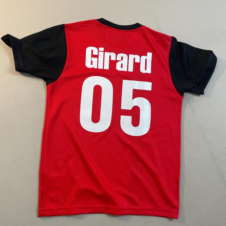 May include: Red jersey with black sleeves and a black collar. The name "Girard" and the number "05" are printed in white on the back of the jersey. The jersey is made of a breathable, athletic material.