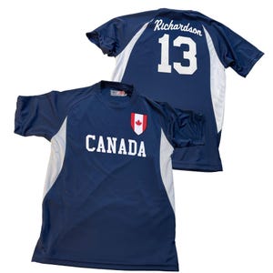 May include: Two navy blue athletic shirts. One shirt has "CANADA" printed in white with a red maple leaf emblem. The other shirt has "Richardson" and the number "13" in white on the back. Both shirts have white side panels.
