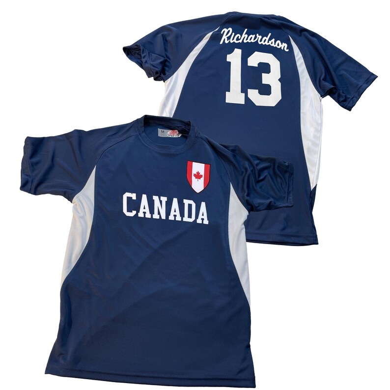 May include: Two navy blue athletic shirts. One shirt has "CANADA" printed in white with a red maple leaf emblem. The other shirt has "Richardson" and the number "13" in white on the back. Both shirts have white side panels.