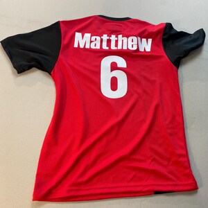 May include: Red athletic jersey with black sleeves. The name "Matthew" and the number "6" are printed in white on the back. The jersey is made of a lightweight, breathable material.