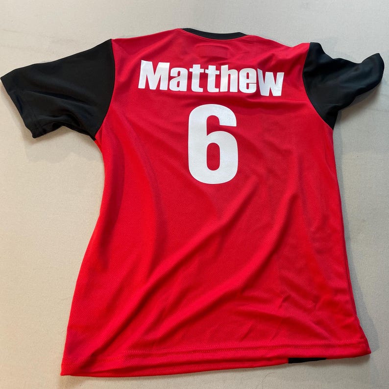 May include: Red athletic jersey with black sleeves. The name "Matthew" and the number "6" are printed in white on the back. The jersey is made of a lightweight, breathable material.