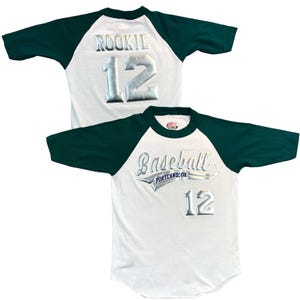 May include: White baseball jersey with dark green sleeves and collar. The jersey features the word "ROOKIE" and the number "12" in silver lettering on the back. The front of the jersey says "Baseball PORTLAND, OR" and the number "12".